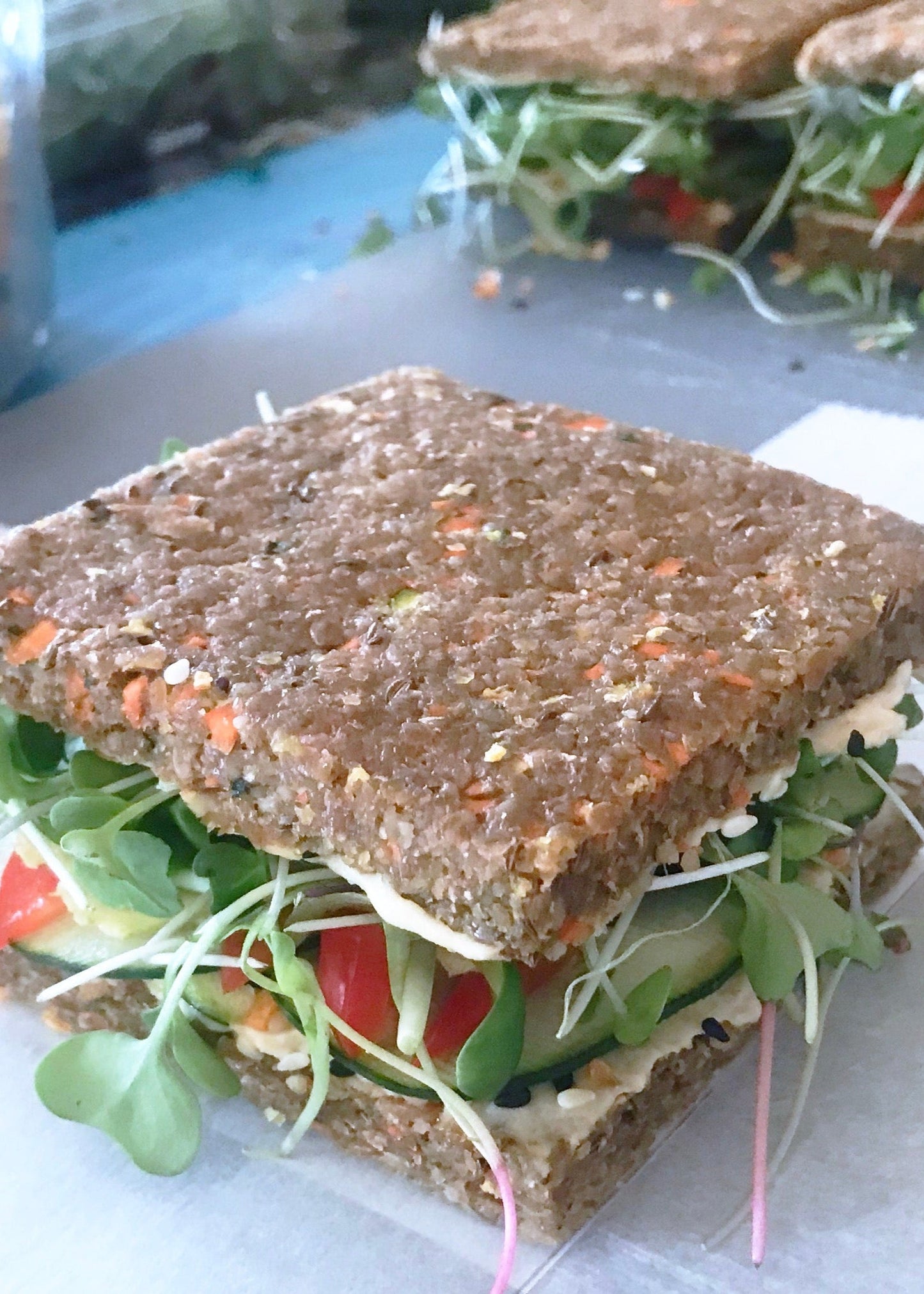 Close-up of a vegetable sandwich made with sprouted raw bread, layered with fresh microgreens, sliced cucumbers, cherry tomatoes, and a spread of hummus.