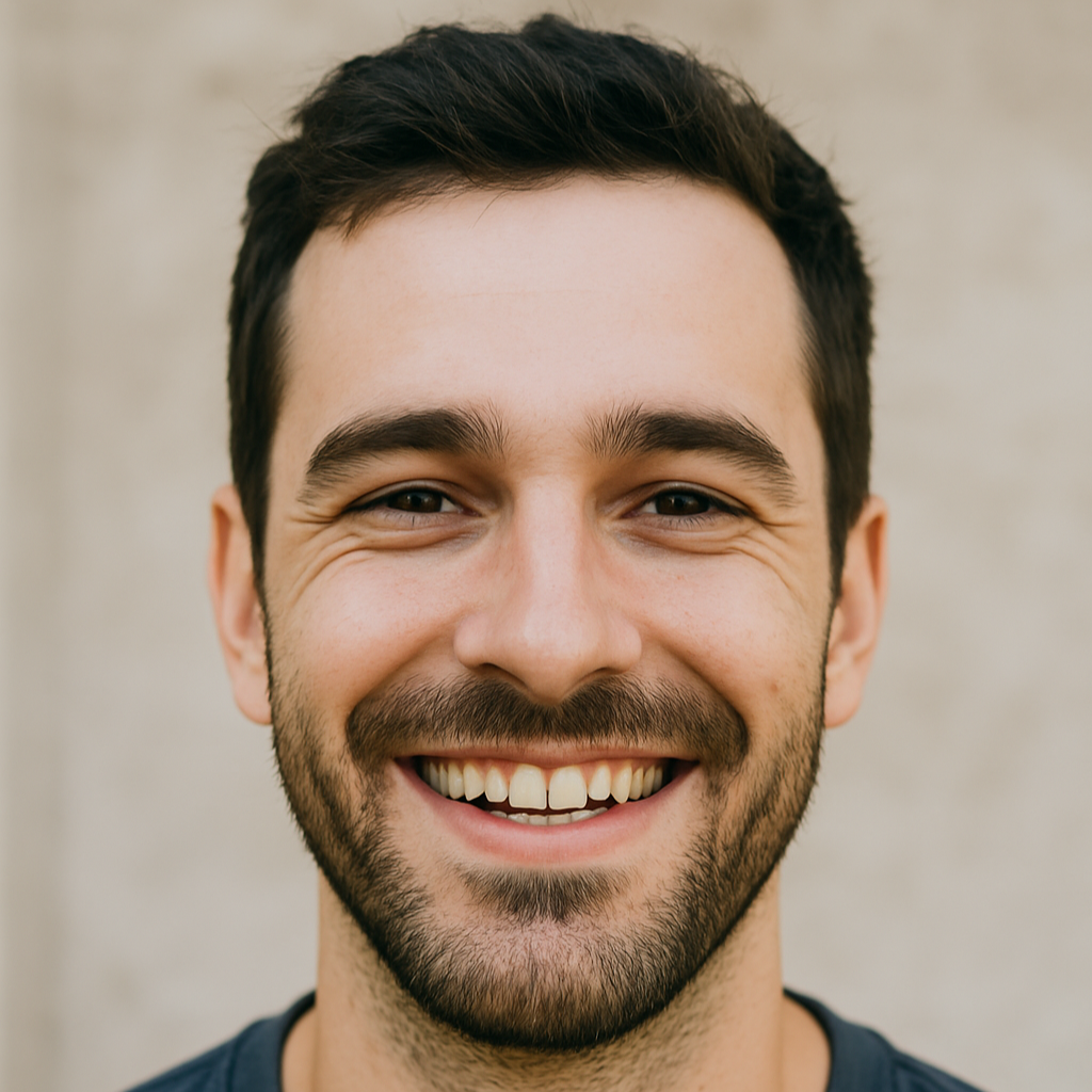 Close-up of a smiling man with short dark hair and a beard, standing against a neutral background.